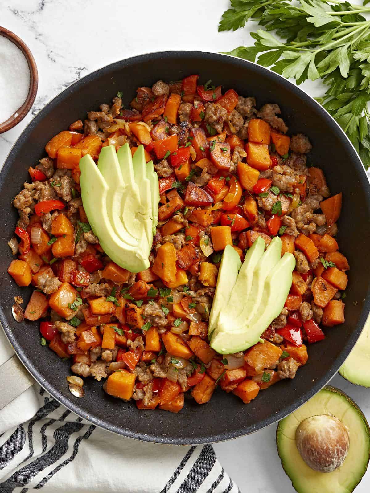 Overhead view of sweet potato hash in a skillet garnished with sliced avocado.