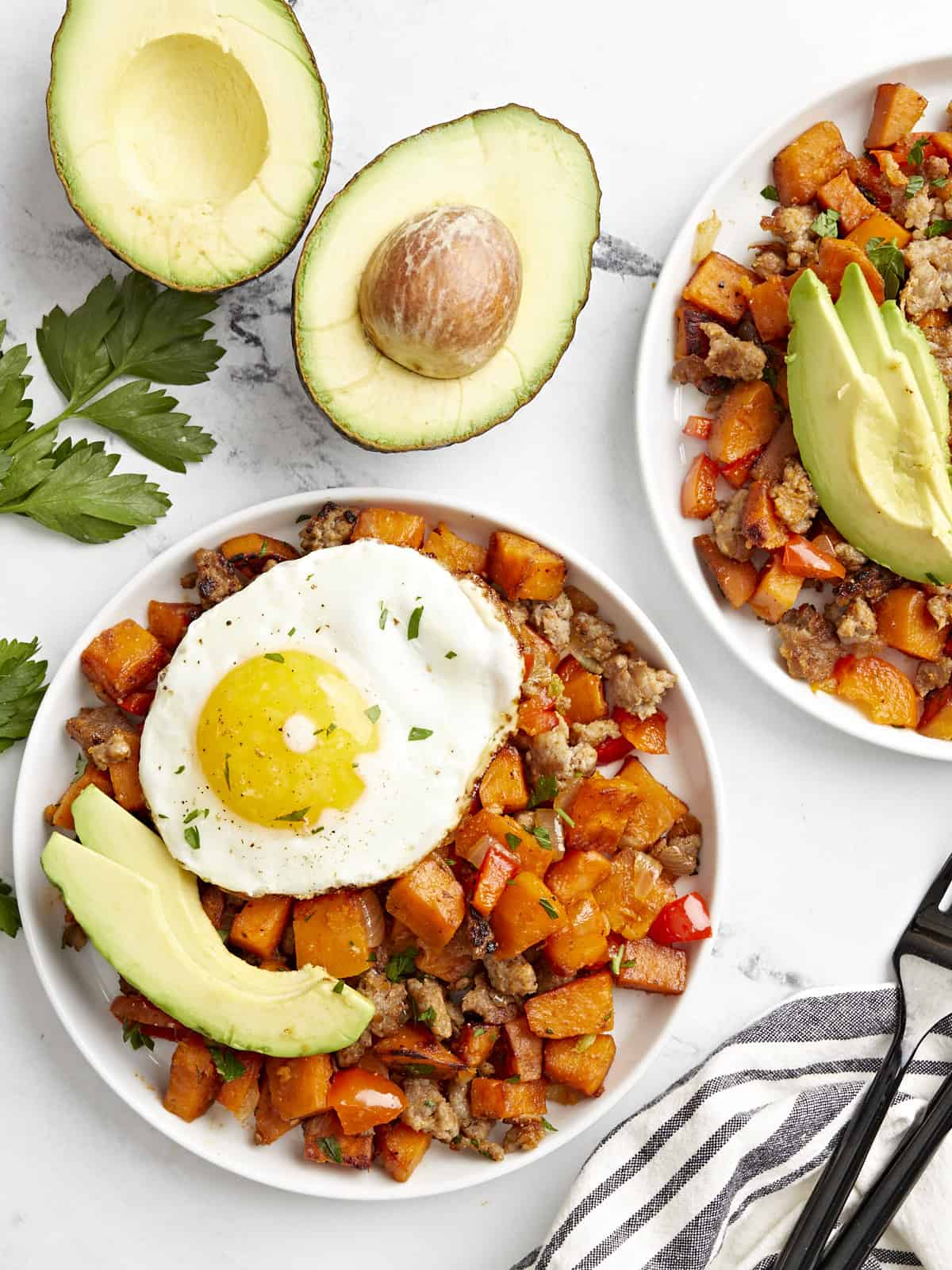 Overhead view of sweet potato hash on two plates topped with sliced avocado and a fried egg.