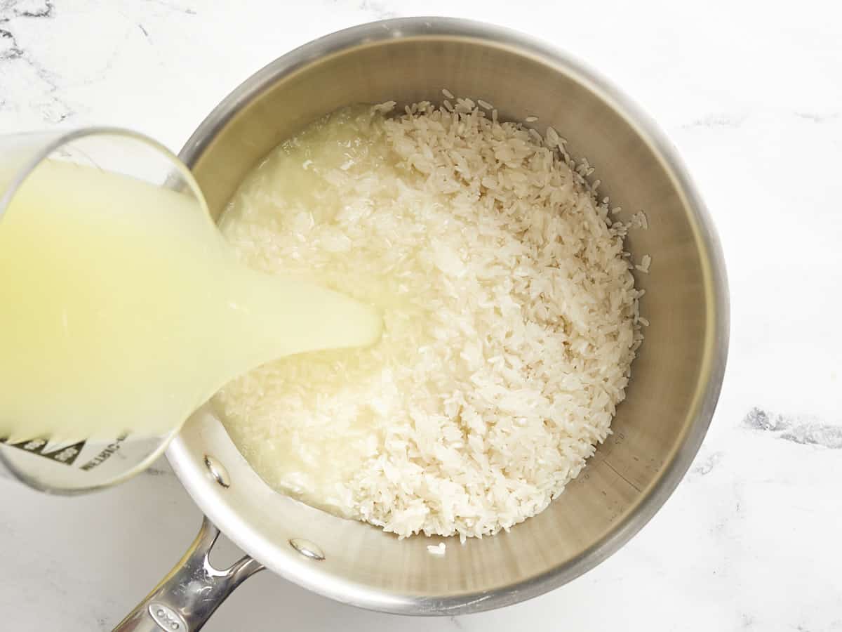 Chicken broth being added to a pot with rinsed rice. 