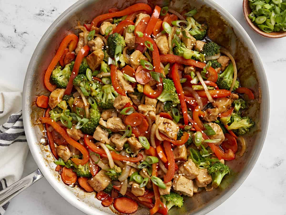 Overhead view of chicken stir fry in a large skillet with sliced green onions on the side.