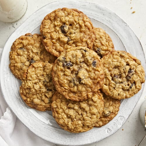 Overhead view of a pile of oatmeal cookies on a plate with a white napkin and a cup of milk on the side.