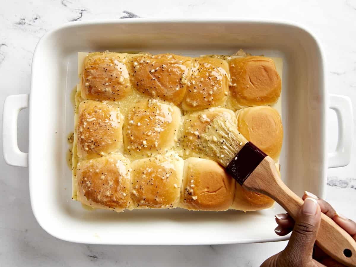 Melted butter sauce being brushed over the tops of hawaiian rolls with a pastry brush.