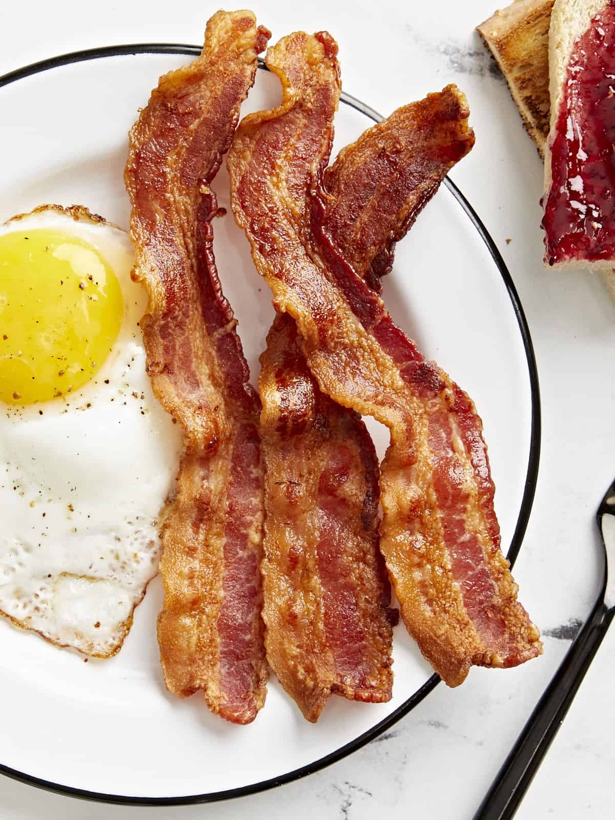 Overhead view of Air Fryer bacon and a fried egg on a white serving plate with toast on the side.