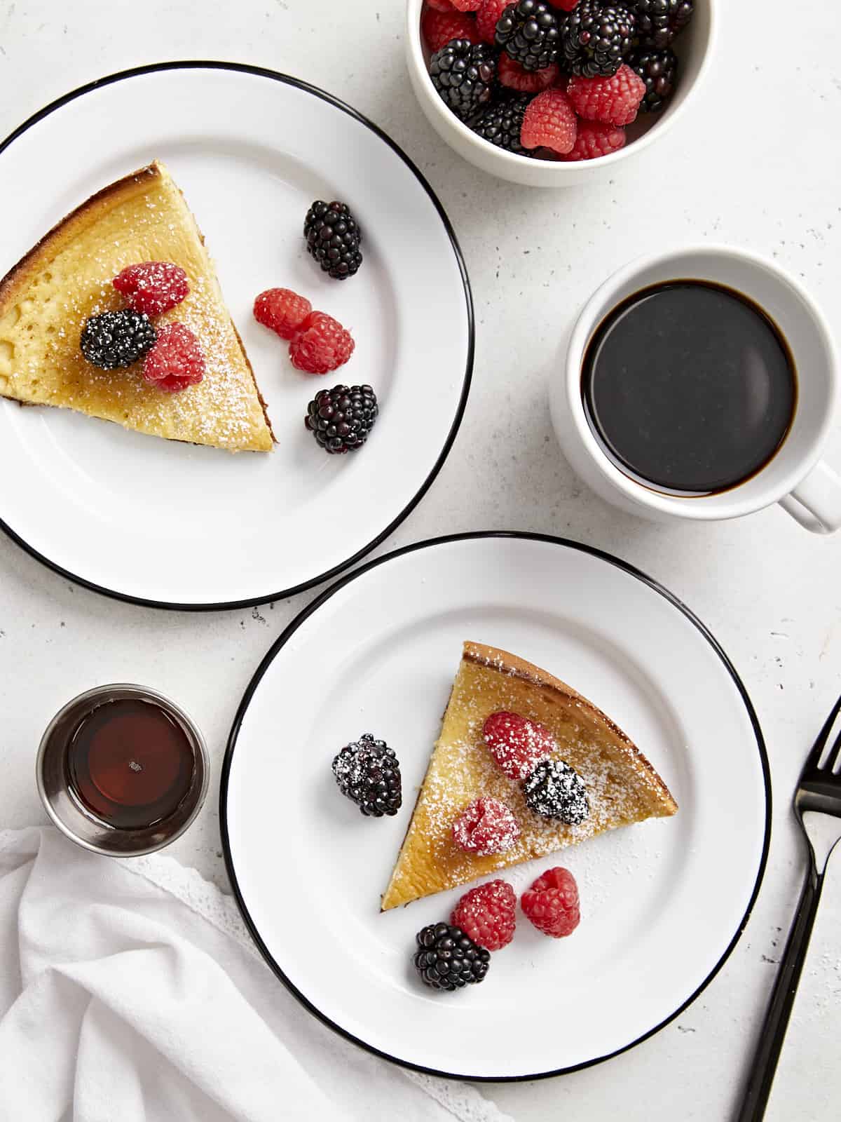 Overhead view of two slices of dutch baby on white serving plates with maple syrup and fresh berries on top.