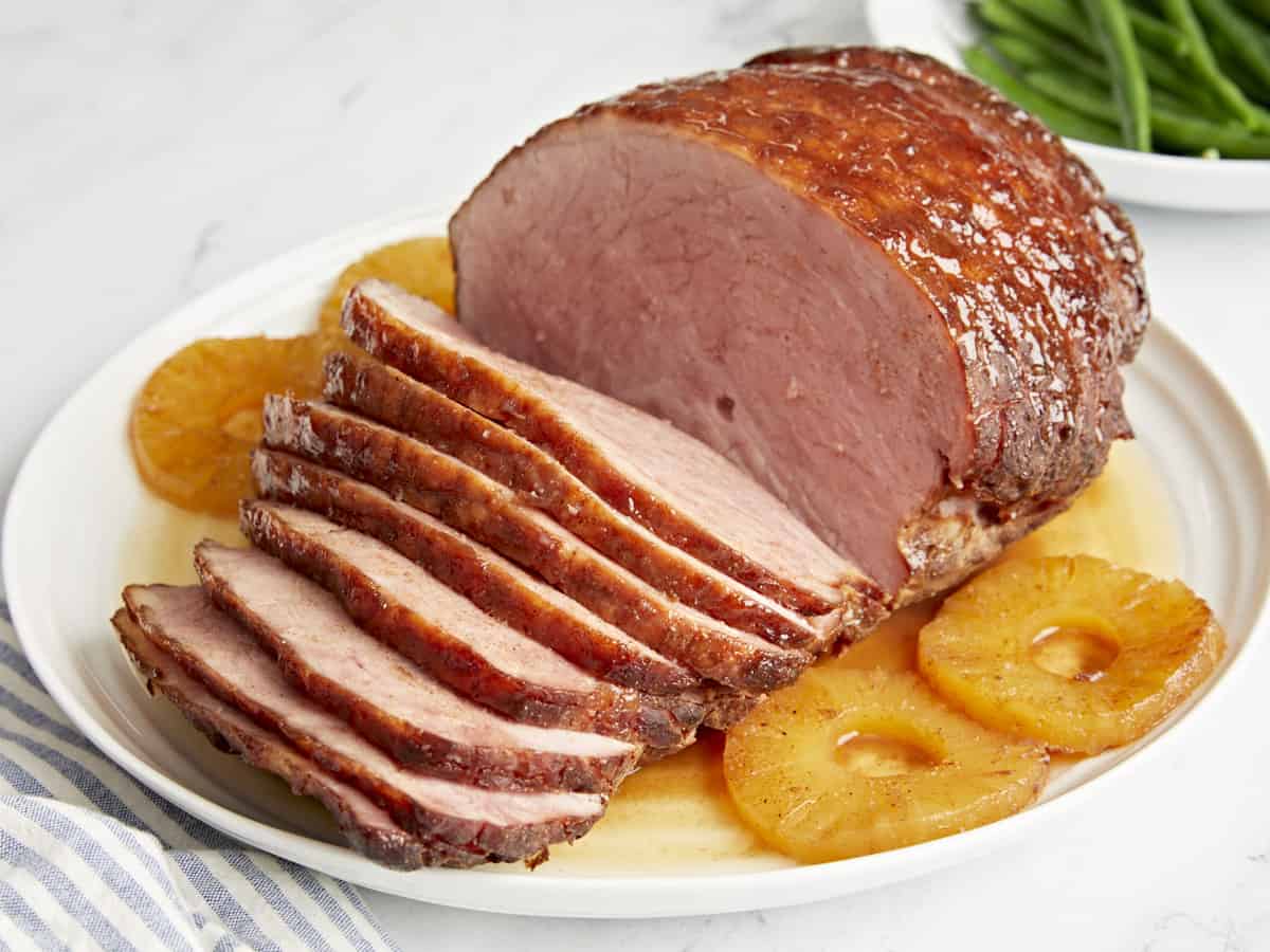 three-quarters view of a partially sliced baked ham on a white plate with pineapple rings.