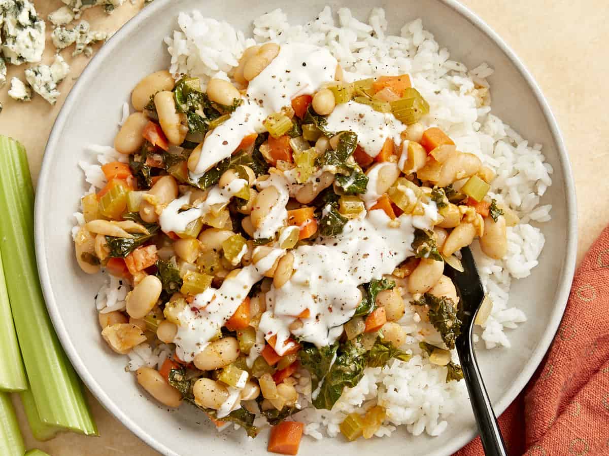 overhead view of buffalo beans and greens over white rice with white sauce on a white plate with a fork.