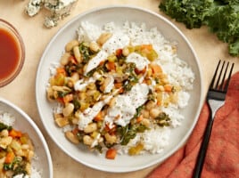 overhead view of buffalo beans and greens over white rice on a white plate with white sauce drizzled over top.