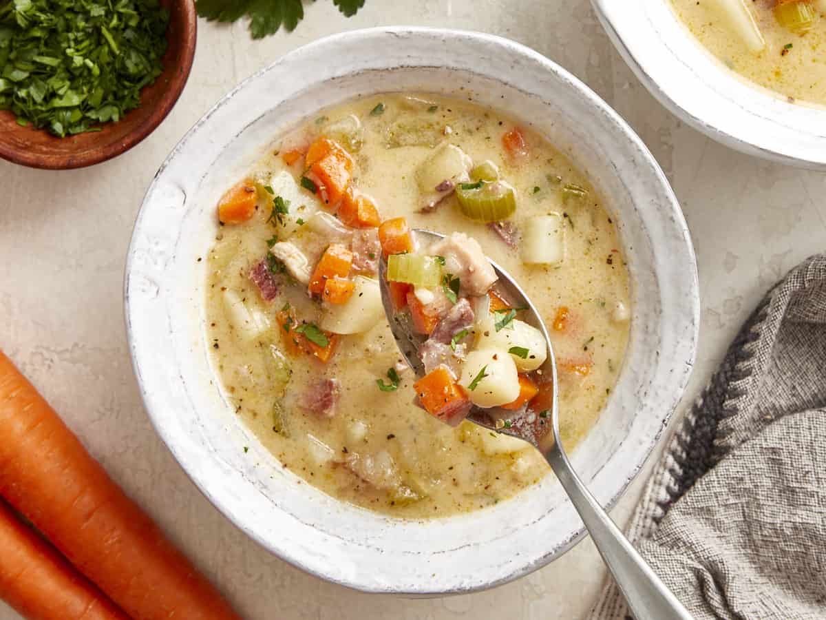 overhead view of a spoonful of ham and potato soup suspended above a serving of soup in a white bowl.