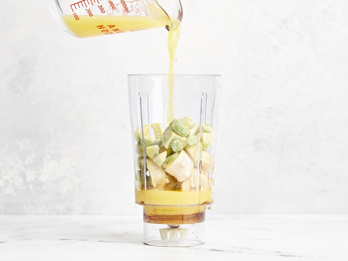 pouring orange juice over ingredients for avocado smoothie in a blender.