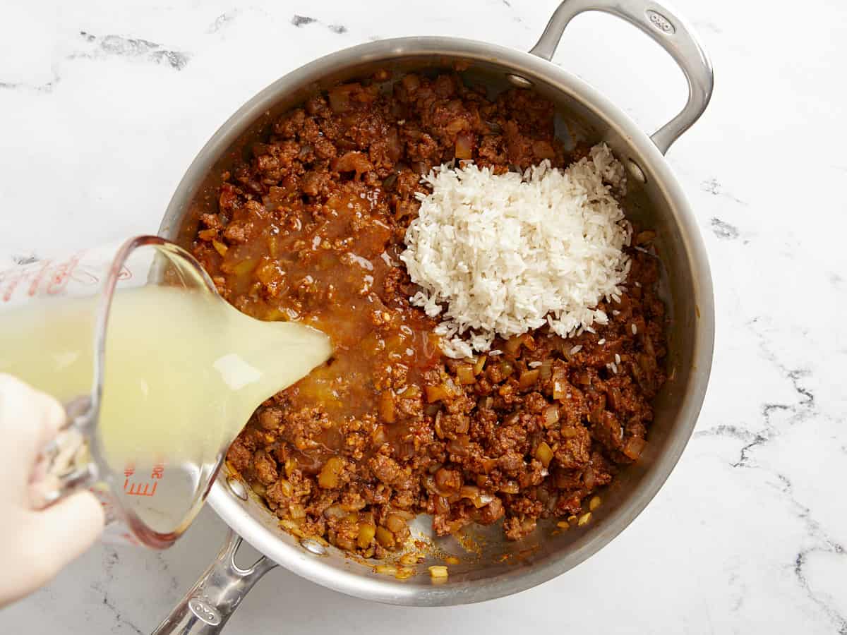 Rice added to the skillet, broth being poured in the side. 