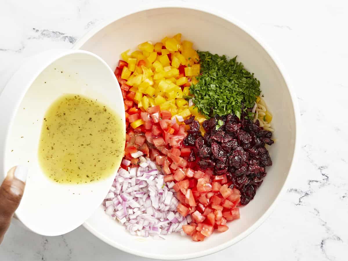 Cooked orzo, corn, and chopped vegetables in a large bowl with vinaigrette dressing being poured on top.