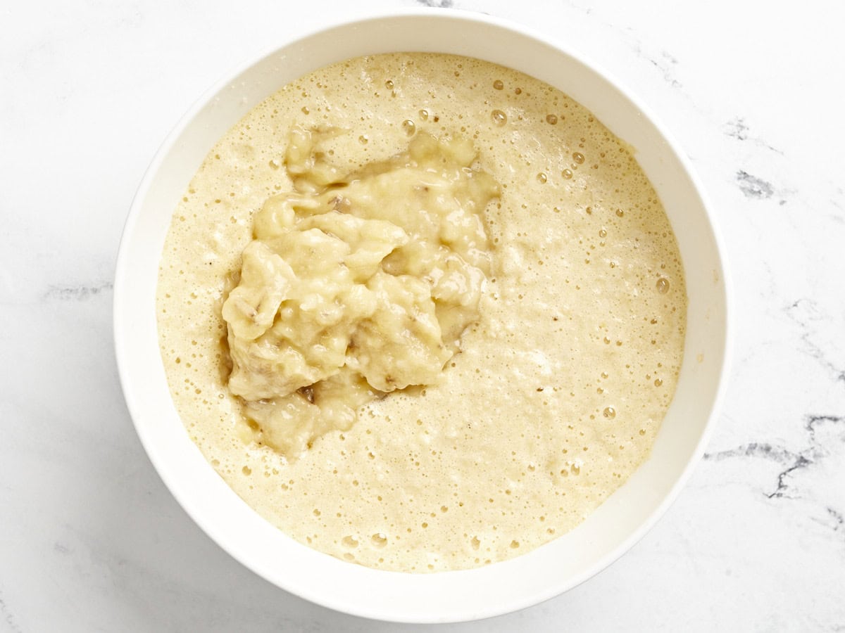 mashed banana added to cake batter in a white bowl.