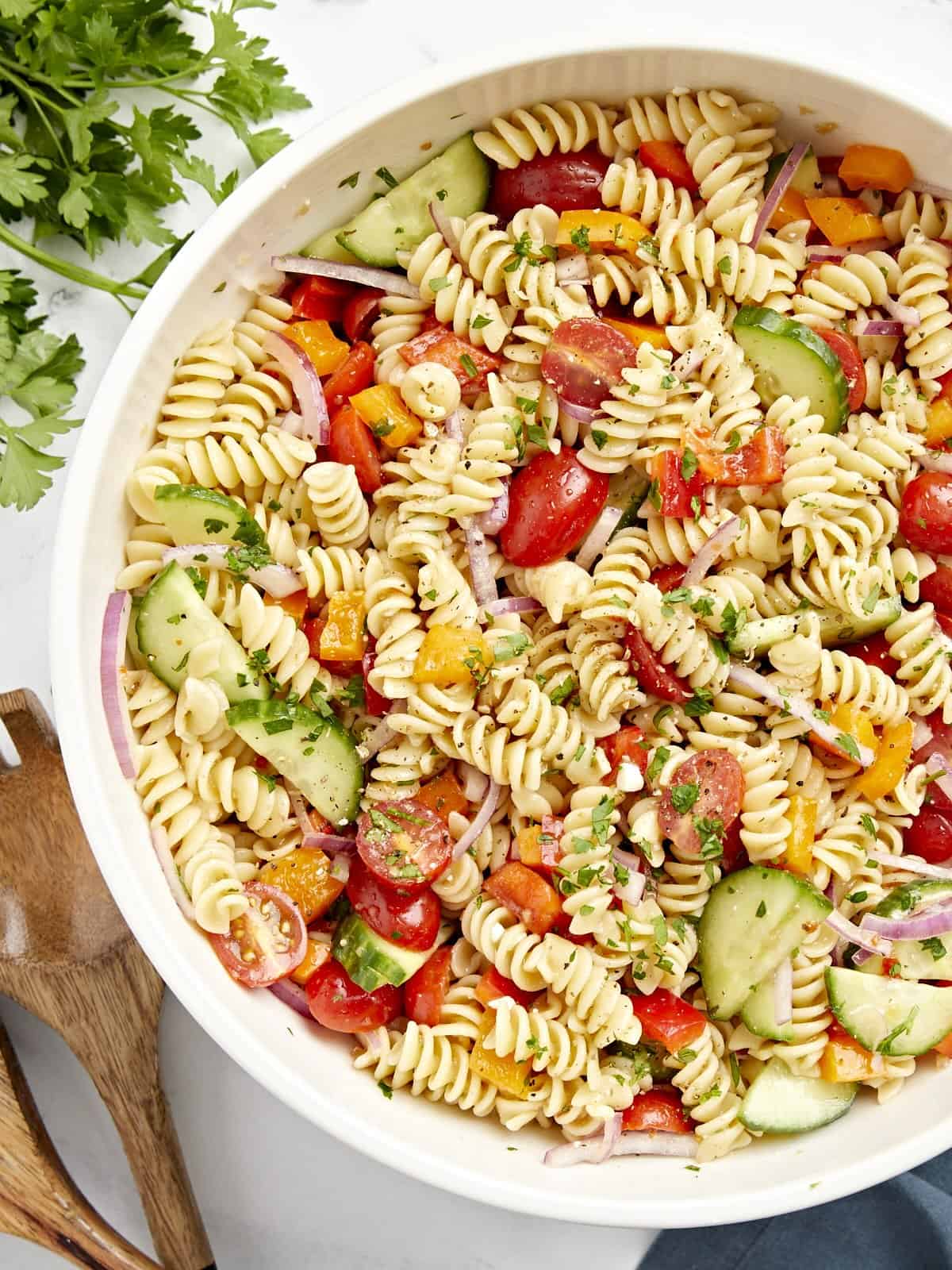 Overhead view of pasta salad in a large white serving bowl with wood serving utensils on the side.