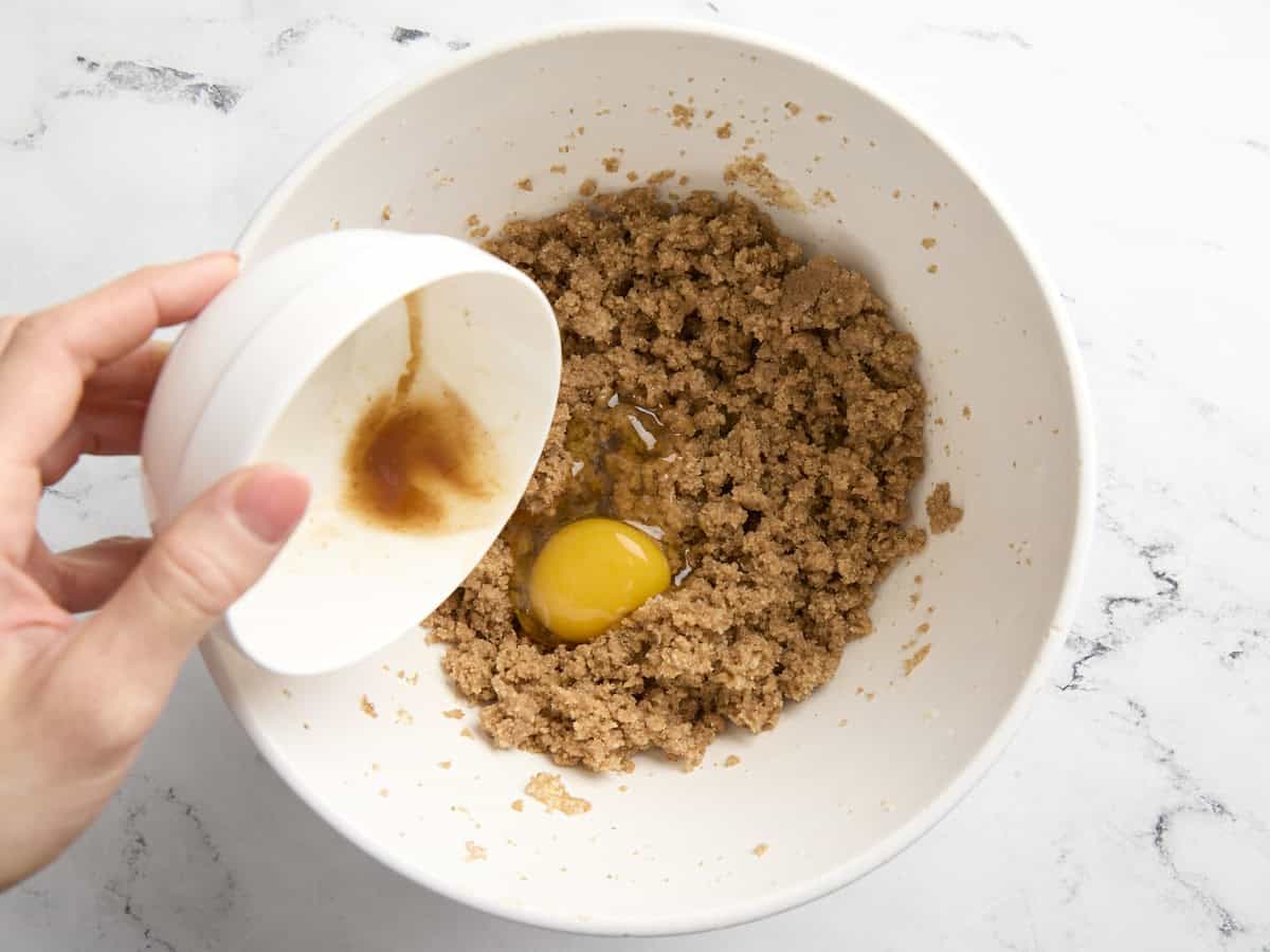 adding an egg and vanilla extract to creamed butter and sugar in a white bowl.