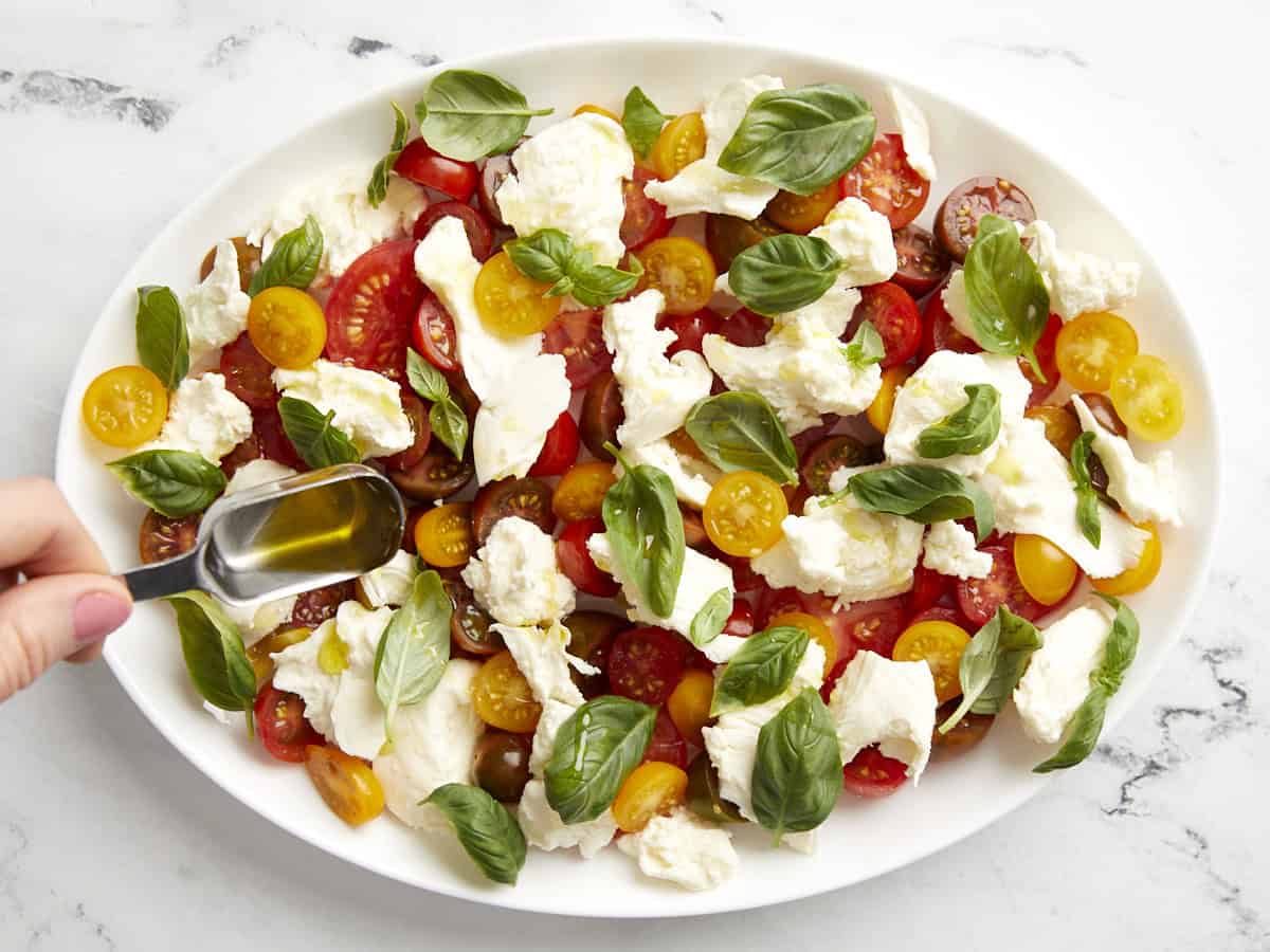 pouring olive oil over caprese salad on a white serving plate.