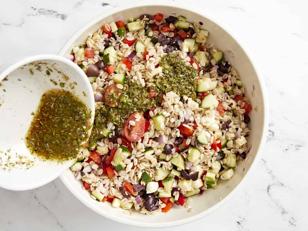 pouring dressing. over barley salad in a white bowl.