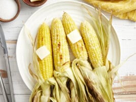 overhead view of 4 peeled ears of grilled corn with butter on a white plate.