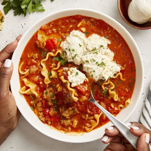 Overhead view of Lasagna Soup in a white bowl with a hand holding a spoon lifting some out.