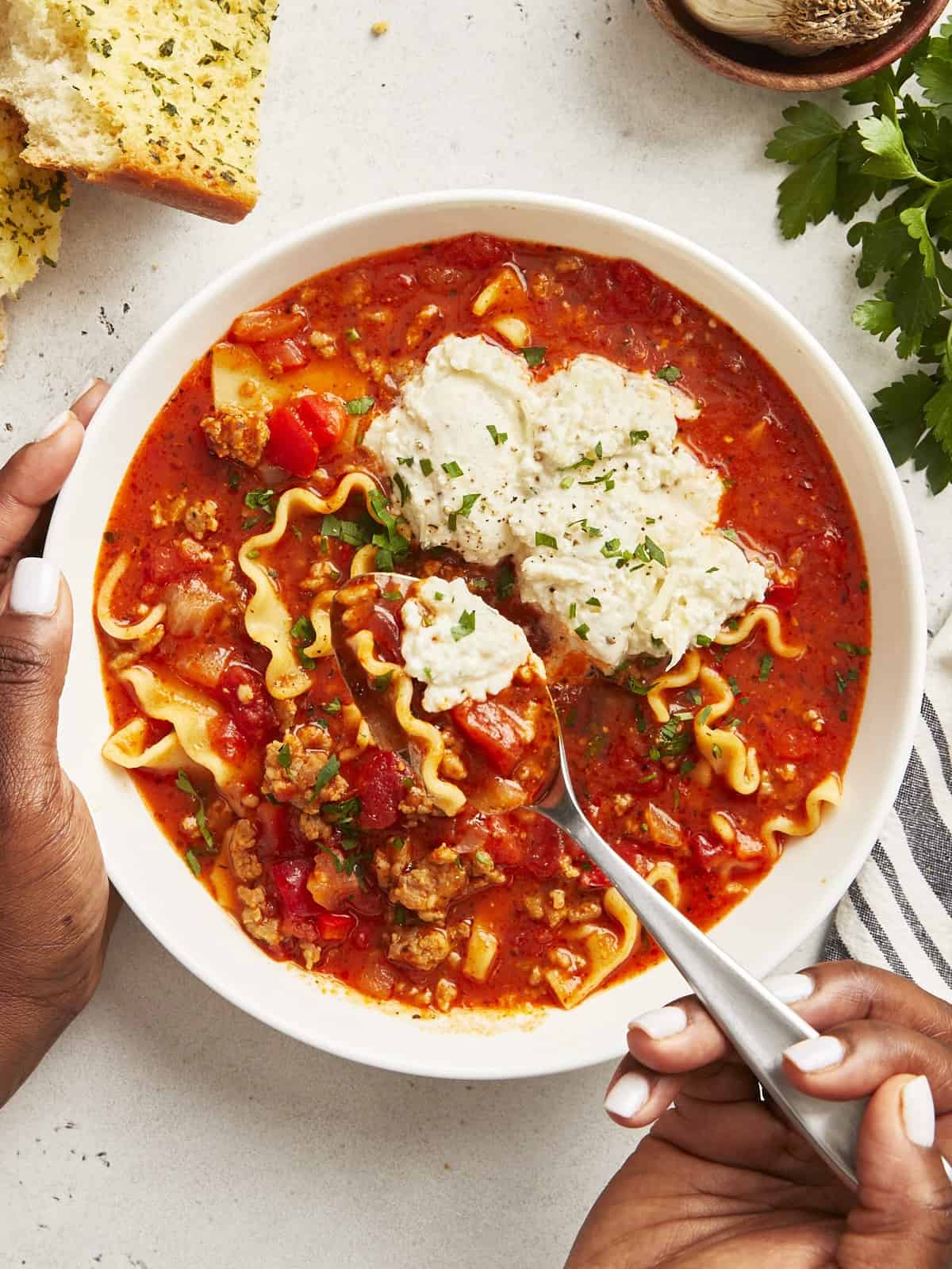 Overhead view of Lasagna Soup in a white bowl with a hand holding a spoon lifting some out.