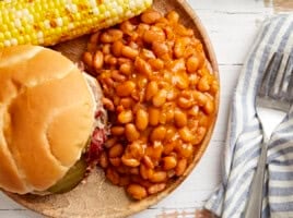 overhead view of a serving of baked beans on a plate with a sandwich and corn on the cob.