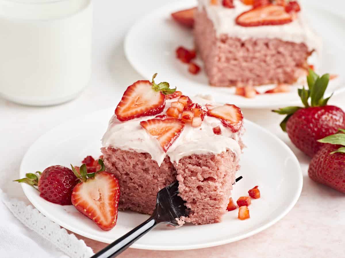 Side view of a slice of strawberry sheet cake on a small white plate with a fork cutting down the middle.