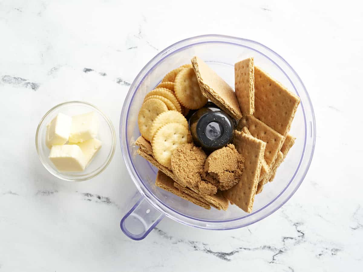 Ritz crackers, brown sugar, and graham crackers in a food processor next to a small bowl of cubed butter.