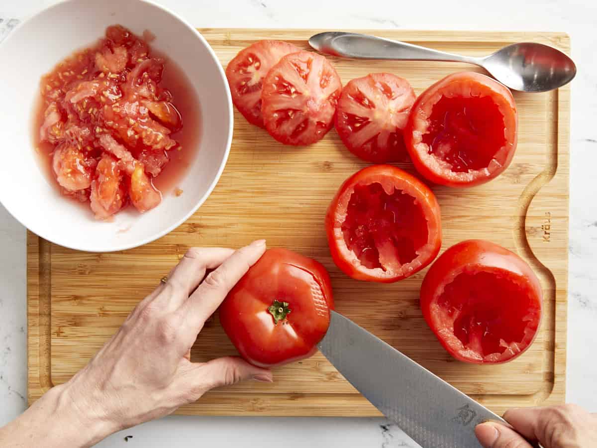 A knife cutting the top off a tomato on a chopping board next to hollowed out tomatoes and a bowl containing scooped out tomato seeds.