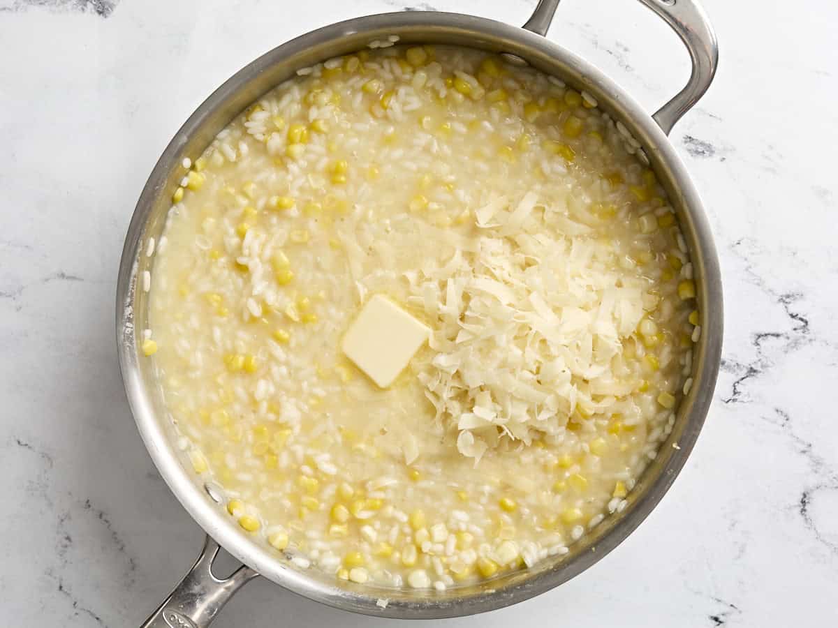 A cube of butter and shredded parmesan cheese added to sweet corn risotto in a skillet.