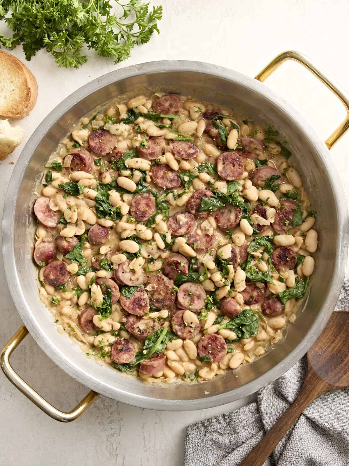 Overhead view of chicken sausage and white beans in a large skillet with a napkin and wooden spoon on the side.