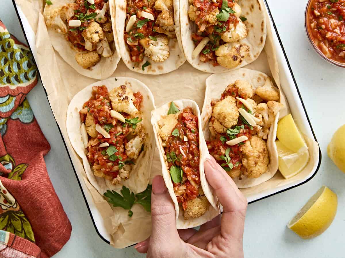 A hand taking a taco from a baking dish full of cauliflower tacos with romesco sauce