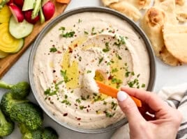 Close up of a hand dipping a carrot stick into a bowl of white bean hummus