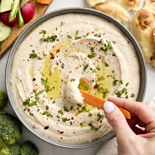 Close up of a hand dipping a carrot stick into a bowl of white bean hummus