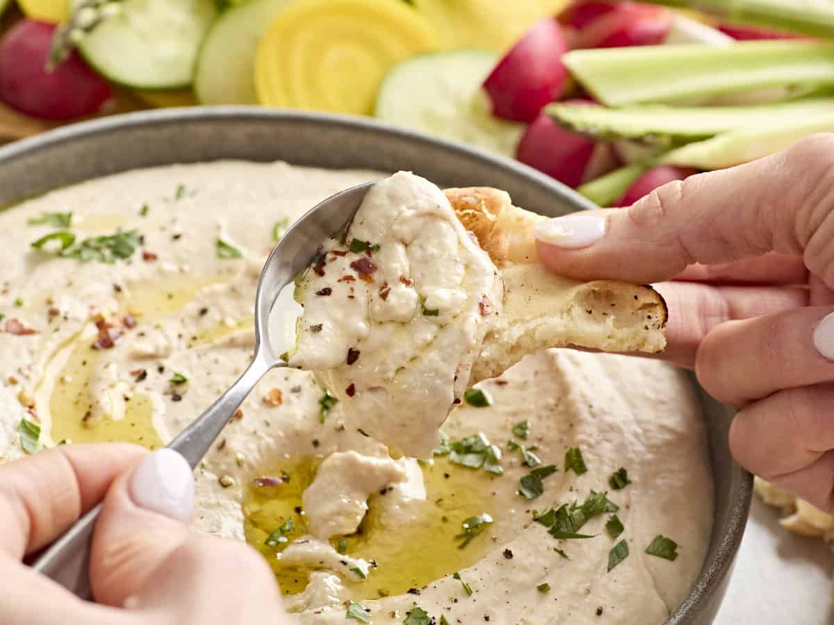 A hand spooning white bean hummus onto pita bread