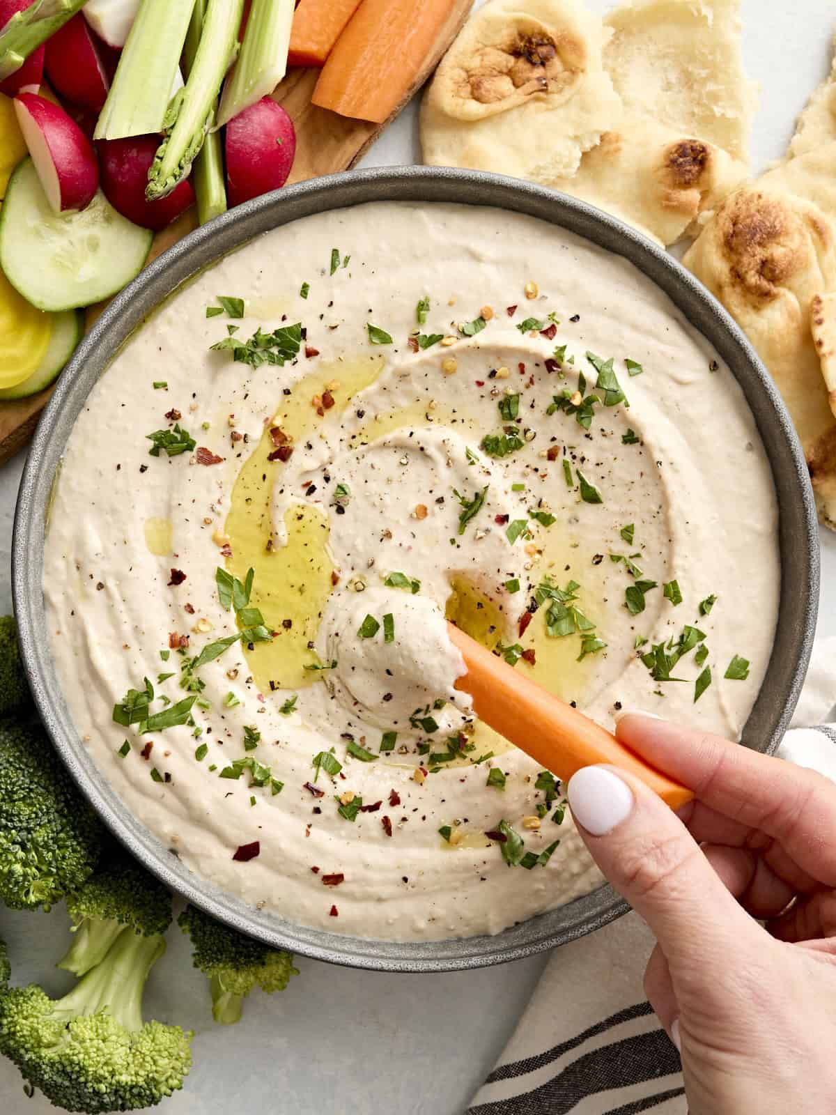 A hand dipping a carrot stick into a bowl of white bean hummus