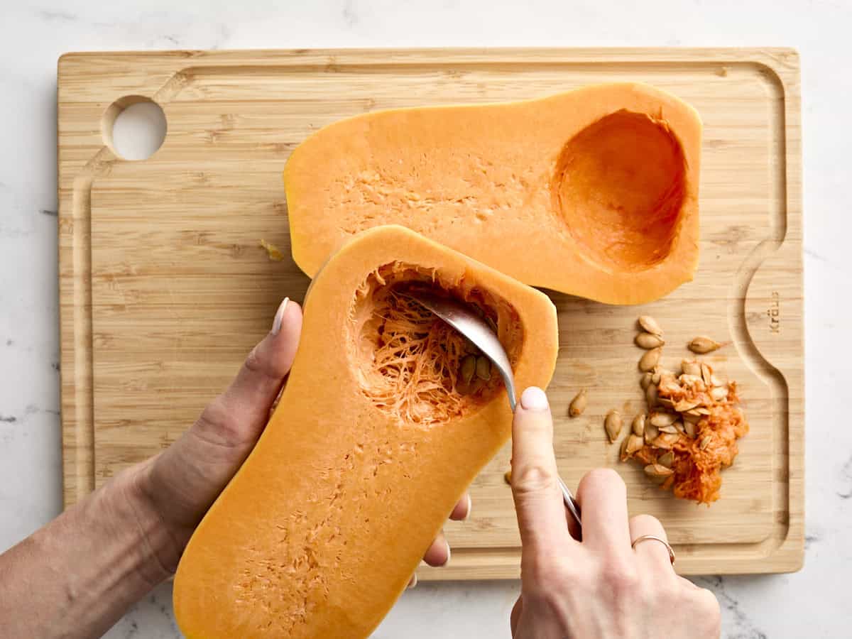 A spoon scooping the stringy membrane and seeds from a butternut squash half.