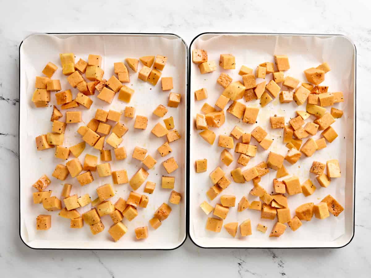 Seasoned butternut squash cubes on two parchment lined baking sheets.