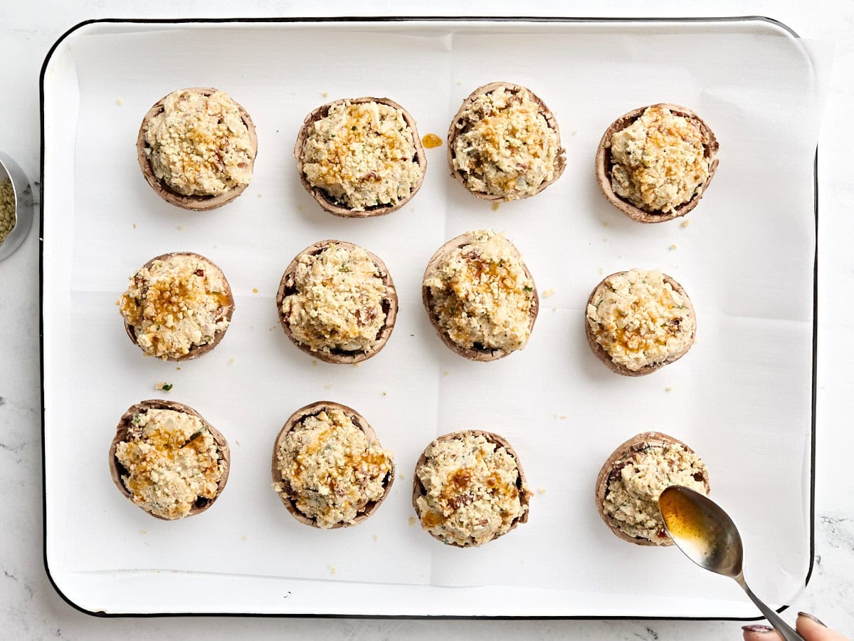 Stuffed mushrooms on a baking sheet with a spoon drizzling oil over.