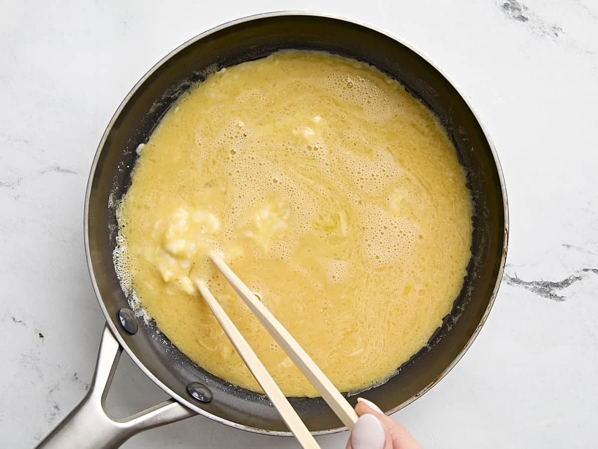 Scrambled egg mixture added to a pan and being whisked by chopsticks.