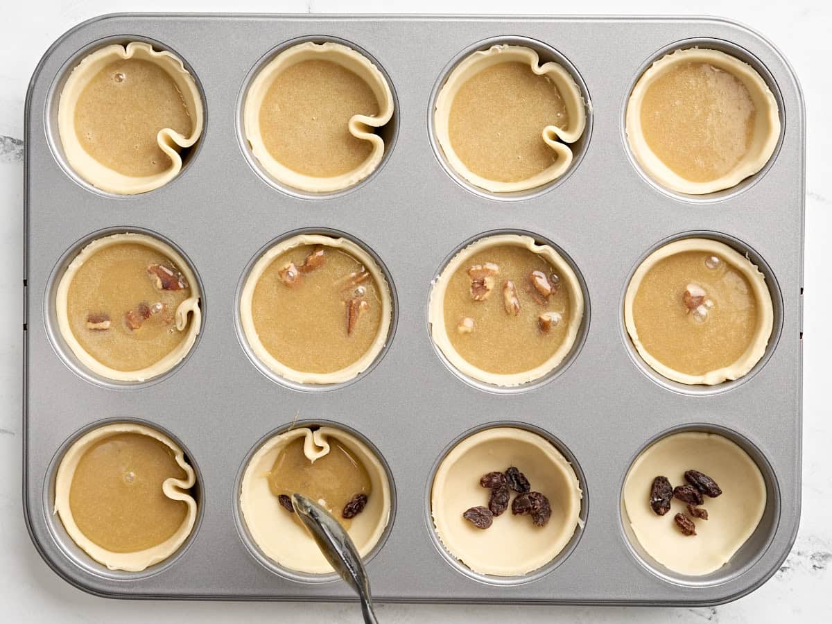 A spoon adding butter tart filling to pie crusts in a cupcake tin, some with pecans and some with raisins.