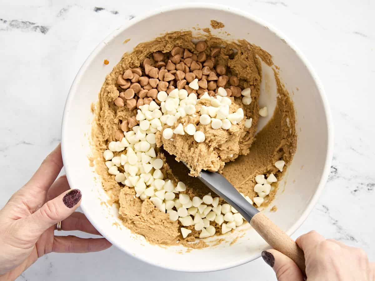 A spatula folding in white chocolate and butterscotch chips into cookie dough.