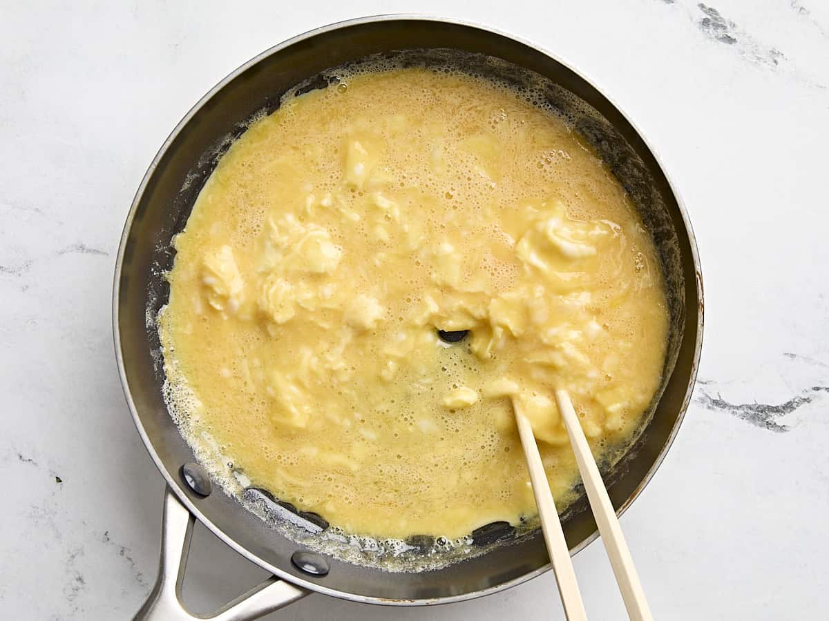 Chopsticks whisking scrambled eggs in a pan.