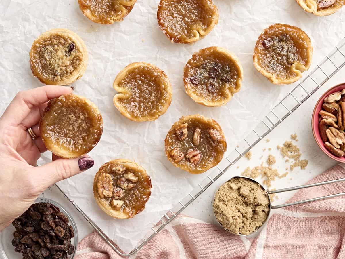 A hand taking a butter tart from a parchment lined cooling rack.