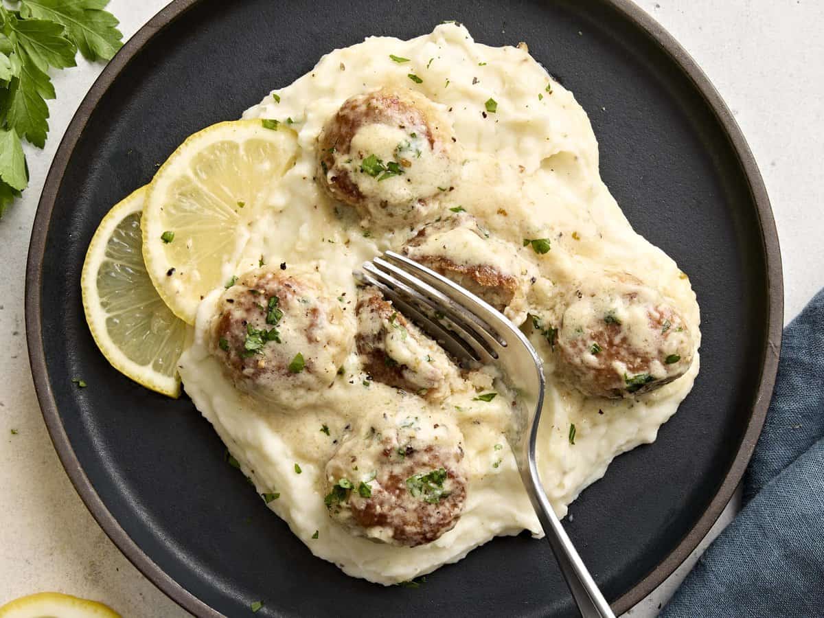 Overhead view of chicken meatballs with a cream sauce served over a plate of mashed potatoes and a fork cutting a meatball in half.