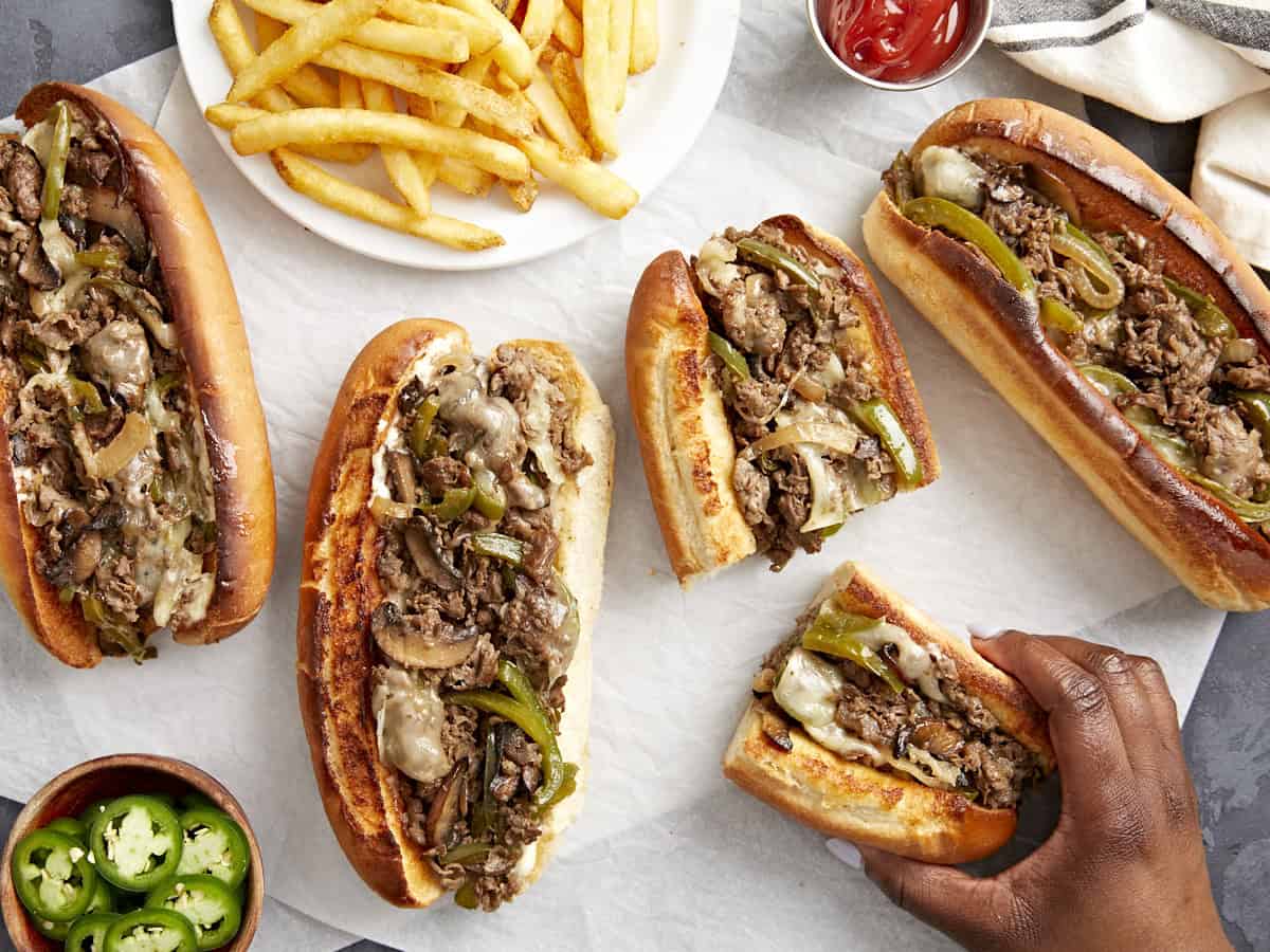 Overhead view of philly cheesesteaks on parchment paper with a hand picking one up.