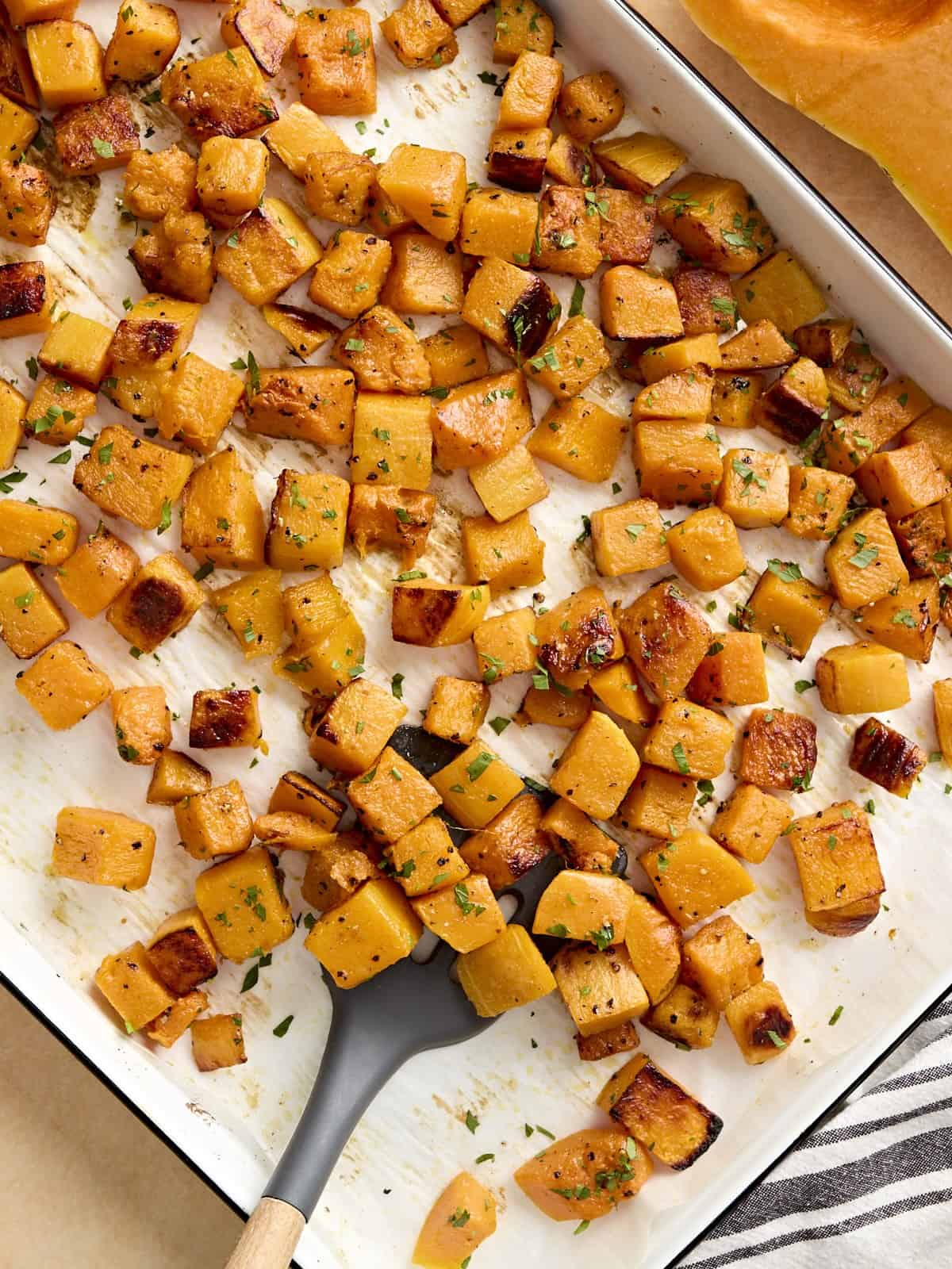 Overhead view of roasted butternut squash cubes on a baking tray with a spatula.