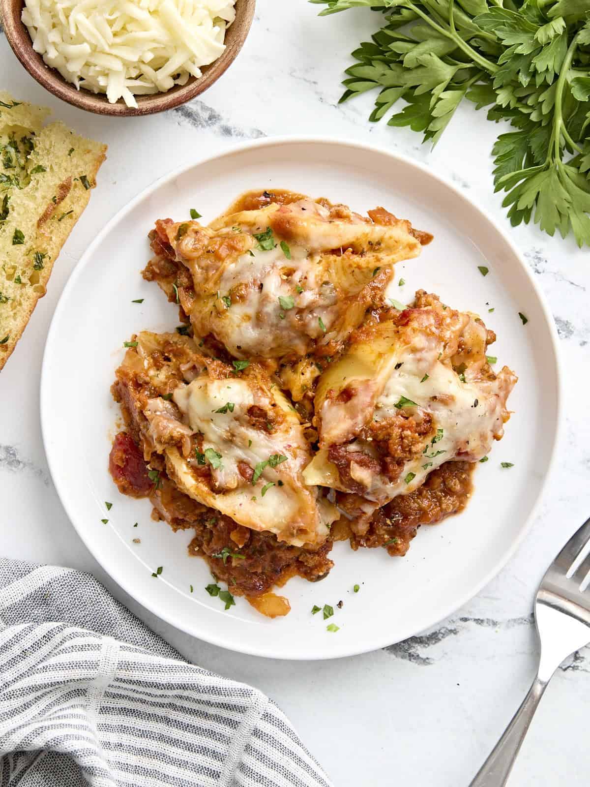 Overhead view of stuffed shells on a white plate with a fork, napkin, and garlic bread on the side.