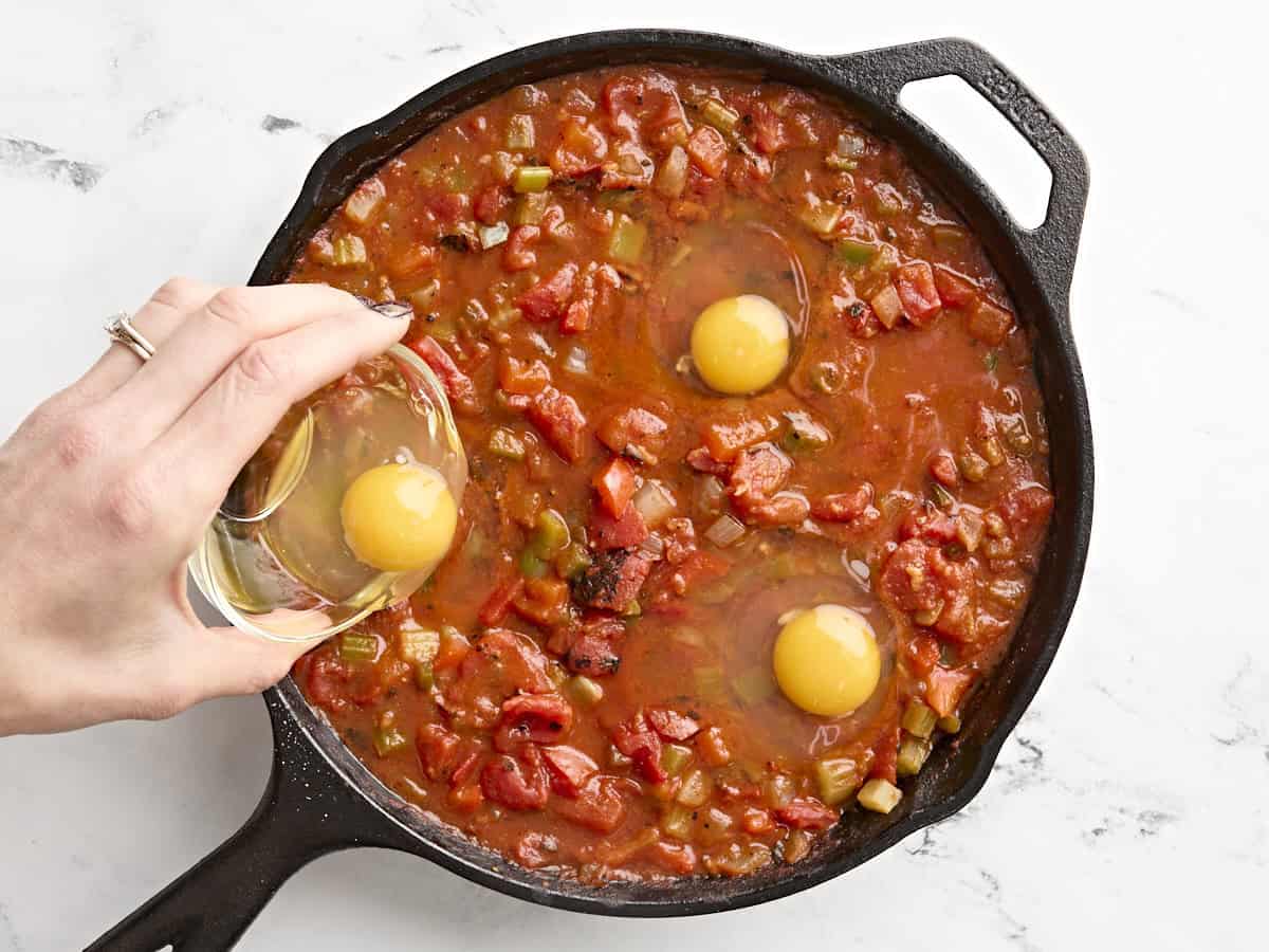 A hand adding an egg to shakshuka in a skillet.