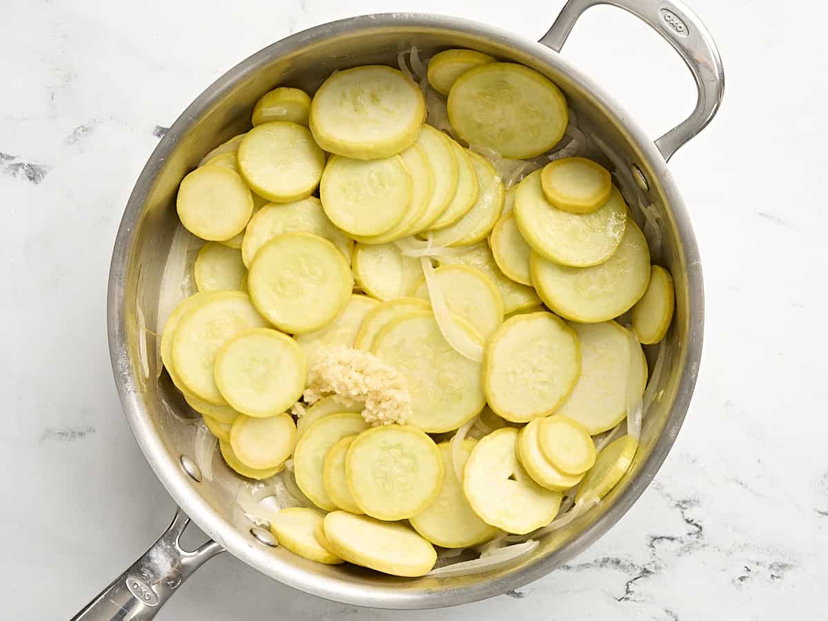 Sliced yellow squash cooking in a skillet with onions and minced garlic.