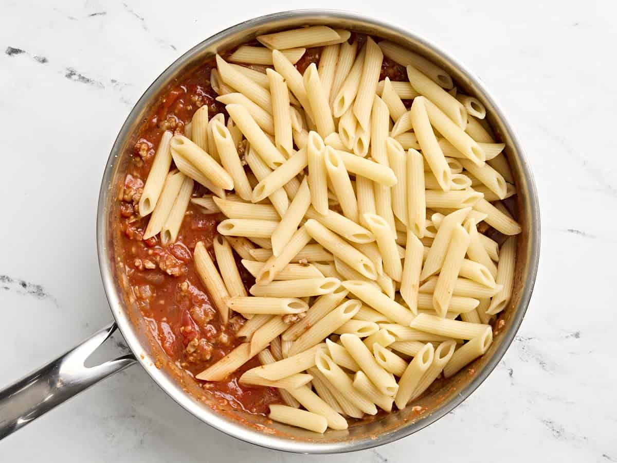 Cooked pasta being added to meat sauce in skillet.