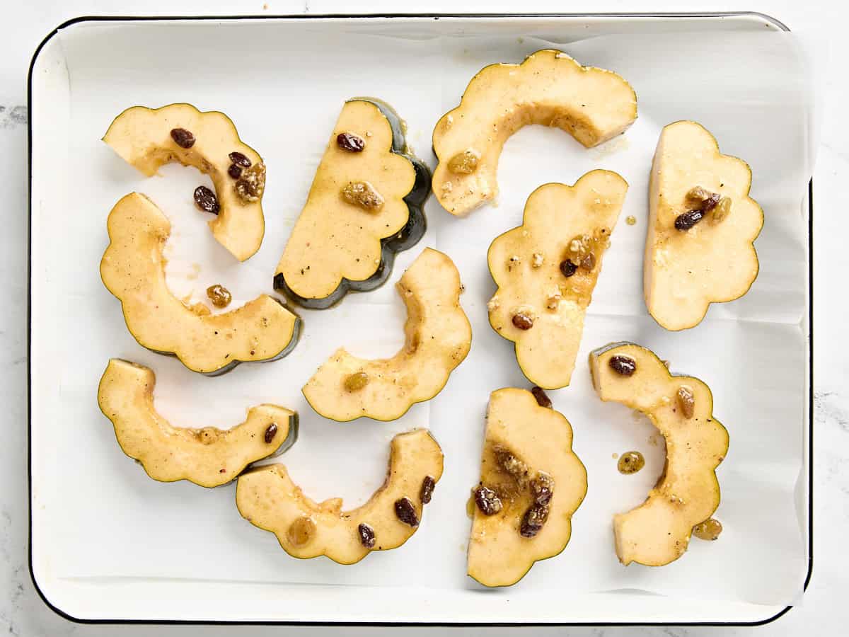 Acorn squash slices coated in oil and raisins on a baking sheet.